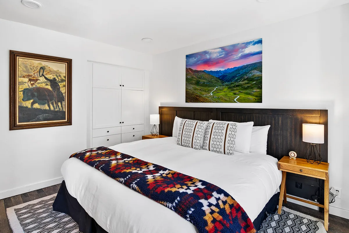 A cozy bedroom with a queen bed draped in a patterned blanket, complemented by warm lighting from bedside lamps and nature-inspired artwork. The inviting setup is ideal for unwinding or enjoying a leisurely morning.