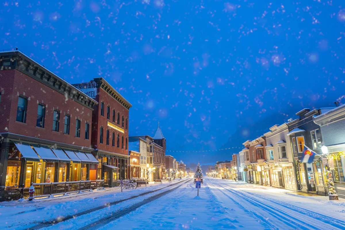 A picturesque Telluride street scene during winter, featuring historic buildings adorned with festive decorations, snow-covered trees, and glowing street lamps. The inviting ambiance hints at local celebrations and a close-knit community atmosphere.