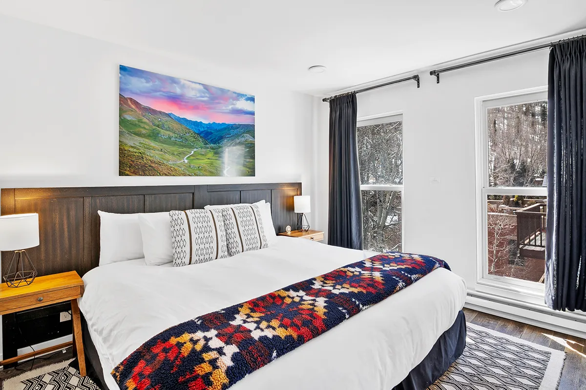 A spacious bedroom featuring a large bed with soft linens, mountain landscape artwork above the headboard, nightstands on each side, and gentle natural light filtering through curtains. The serene atmosphere is conducive to relaxation and restful sleep.