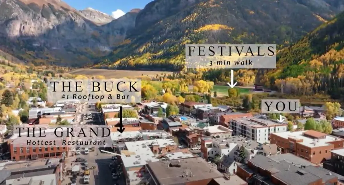 A lively town square set against a backdrop of majestic mountains and vibrant fall foliage. Rooftop bars, restaurants, and local shops line the streets, and people can be seen enjoying outdoor dining and exploring the area. The scene is energized with the buzz of local festivals and scenic walks in the surrounding nature.