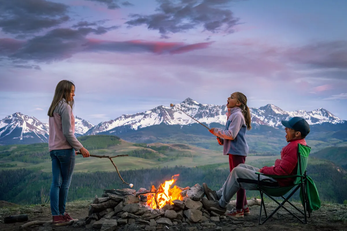 A cozy family scene outdoors with children roasting marshmallows over a campfire, mountains in the background, and parents nearby. The setting is playful and warm, emphasizing togetherness and creating cherished memories in nature.