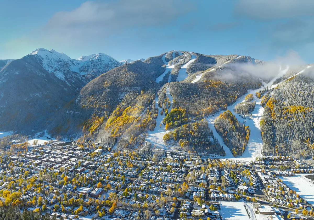 An elevated perspective showing the charming town of Telluride nestled below snow-capped mountains, colorful autumn trees, and ski slopes. The image blends the vibrant energy of the town with the majestic mountain landscape, illustrating both adventure and walkable convenience.
