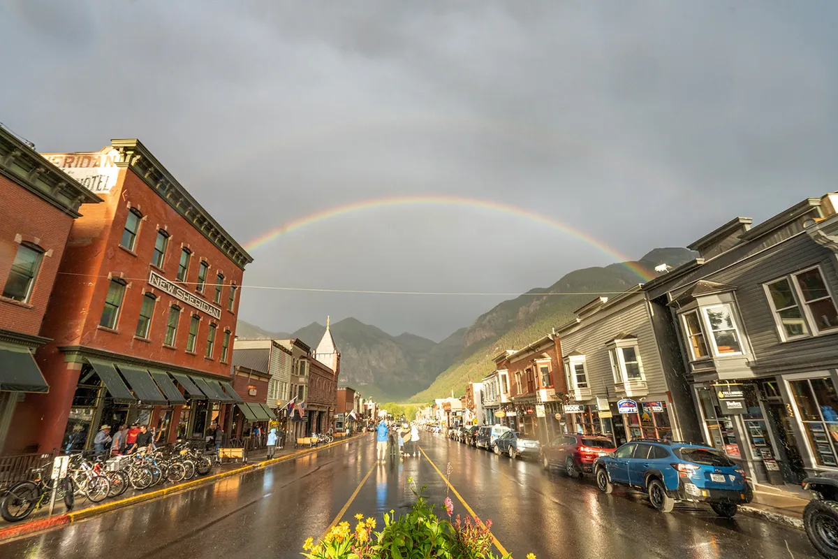 A vibrant Telluride street scene featuring historic buildings, a rainbow arcing over wet pavement, bicycles, and people walking. The image captures an active town ambiance with locals and visitors exploring shops and cafes.