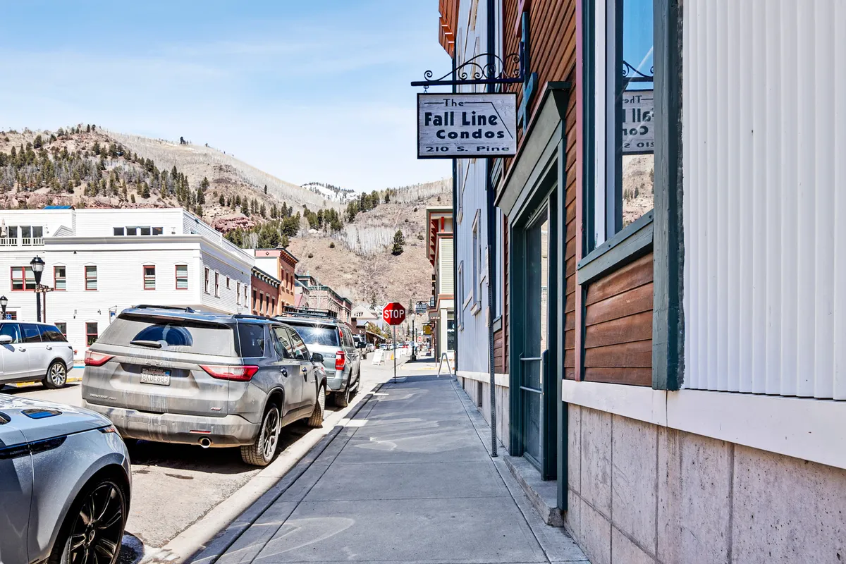 The exterior shot features the Fall Line Condos sign, mountain views in the background, a vibrant street scene, and parked cars, indicating easy access and a central location in Telluride.