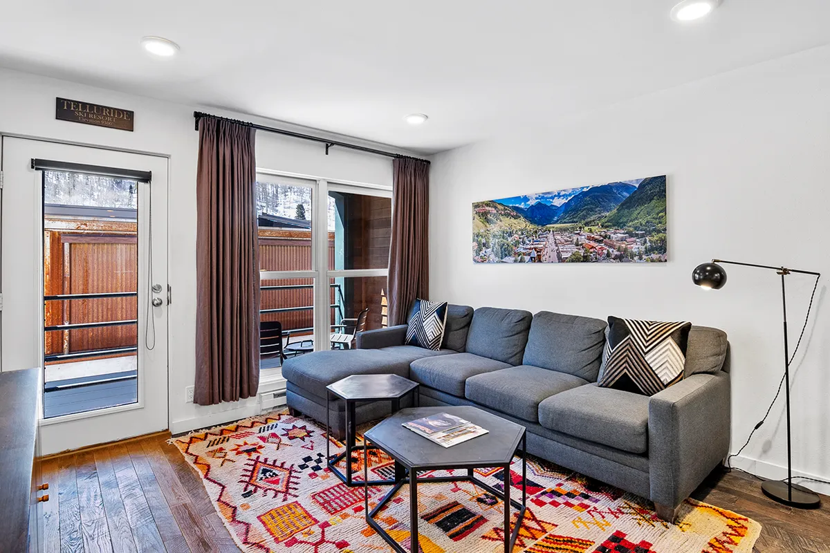 The image showcases a stylish, sunlit living area with a cozy sofa, inviting rug, tasteful artwork, and large windows that flood the room with natural light. The windows offer glimpses of mountain views, creating a welcoming space perfect for relaxing, socializing, or enjoying a peaceful morning coffee.