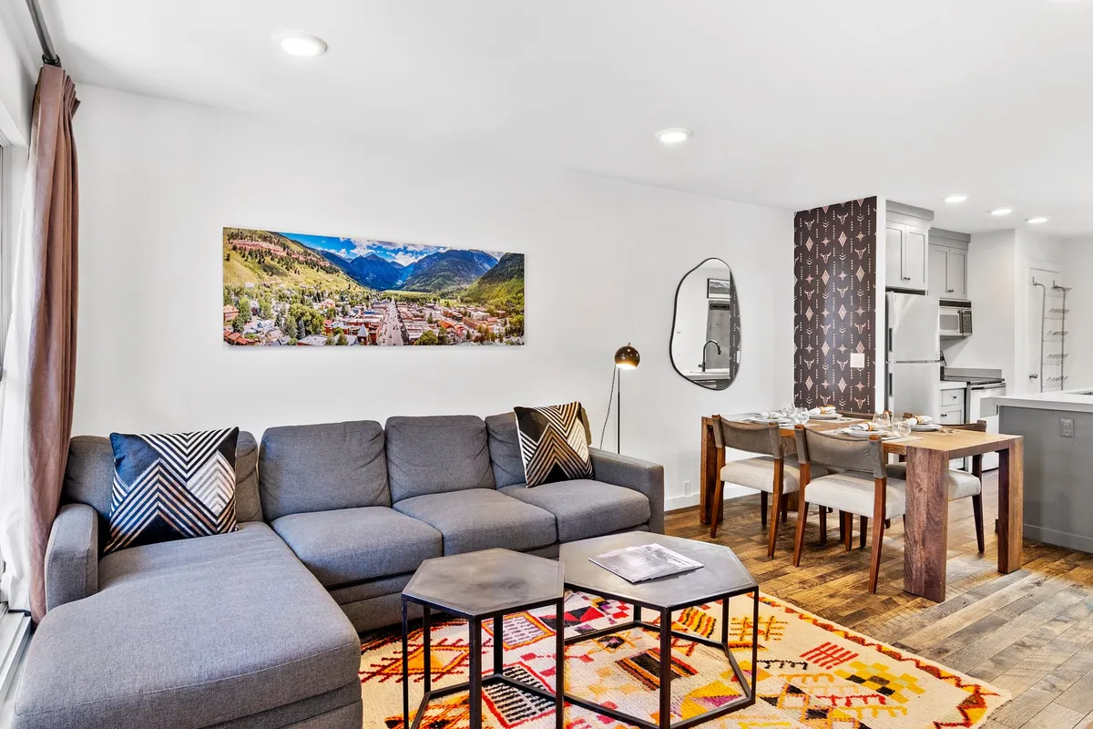 This image showcases a stylish, contemporary living and dining area. The focal points include a plush sofa positioned for relaxing, a dining table set for a gathering, modern artwork on the wall, and a cozy rug anchoring the space. The natural light enhances the room's welcoming atmosphere, highlighting the open-plan arrangement.