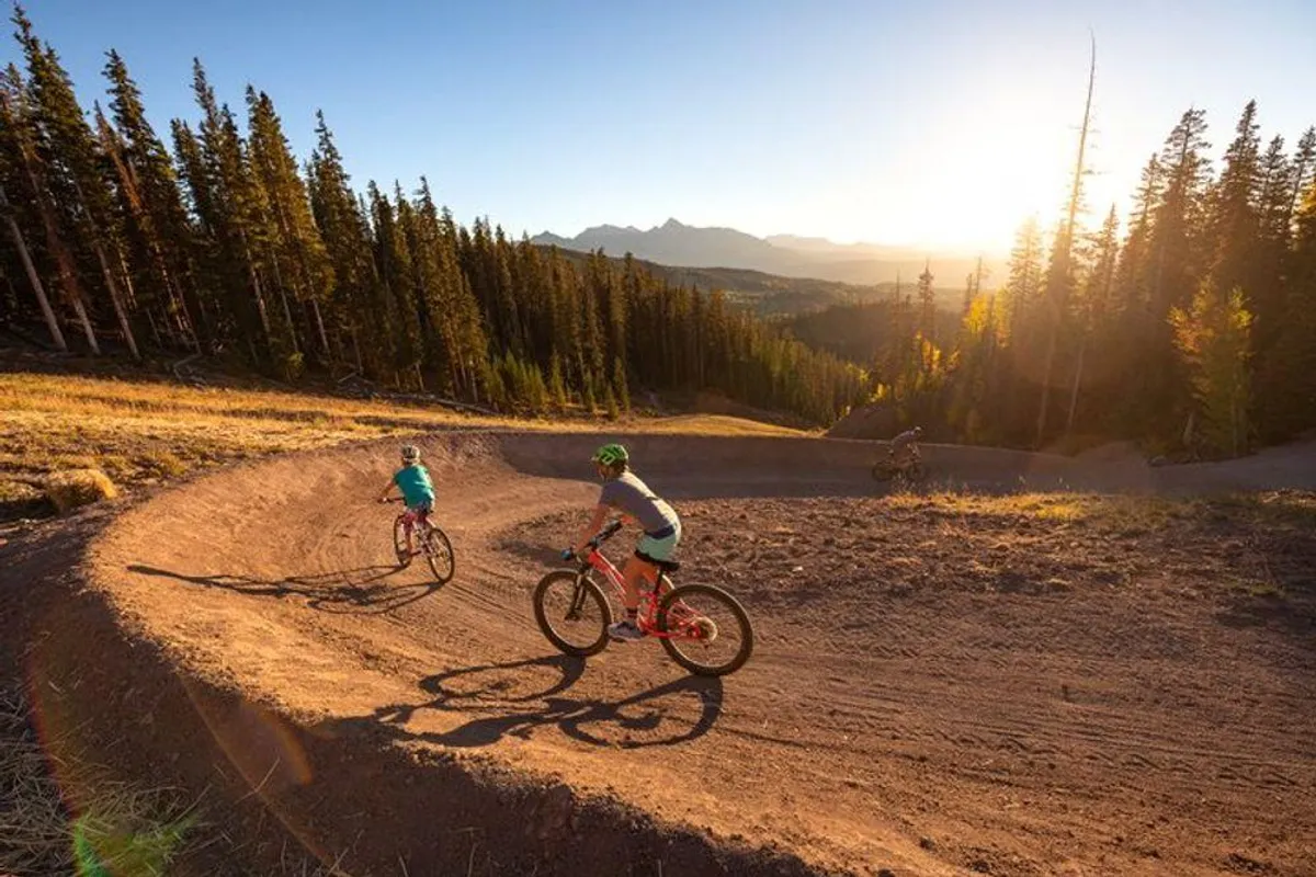 A scenic mountain path winds through lush greenery, with bicyclists riding along and expansive mountain peaks in the background. The image evokes the thrill of outdoor activities like hiking and mountain biking amid spectacular natural beauty.